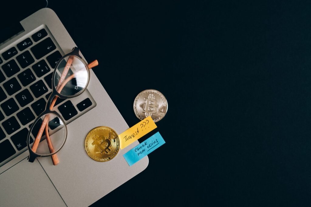 Bitcoin coins, reading glasses and handwritten notes on a desk beside a laptop, representing crypto record-keeping and reconciliation